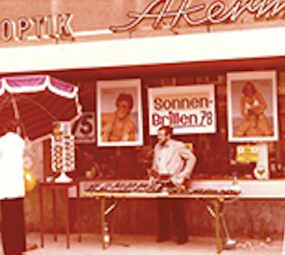 Verkäufer an einem Stand vor Optik-Laden mit Sonnenbrillen und Schildern aus den 70ern.