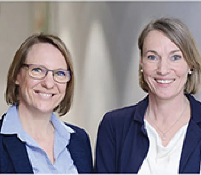 Zwei lächelnde Frauen in Businesskleidung vor einem unscharfen Hintergrund.