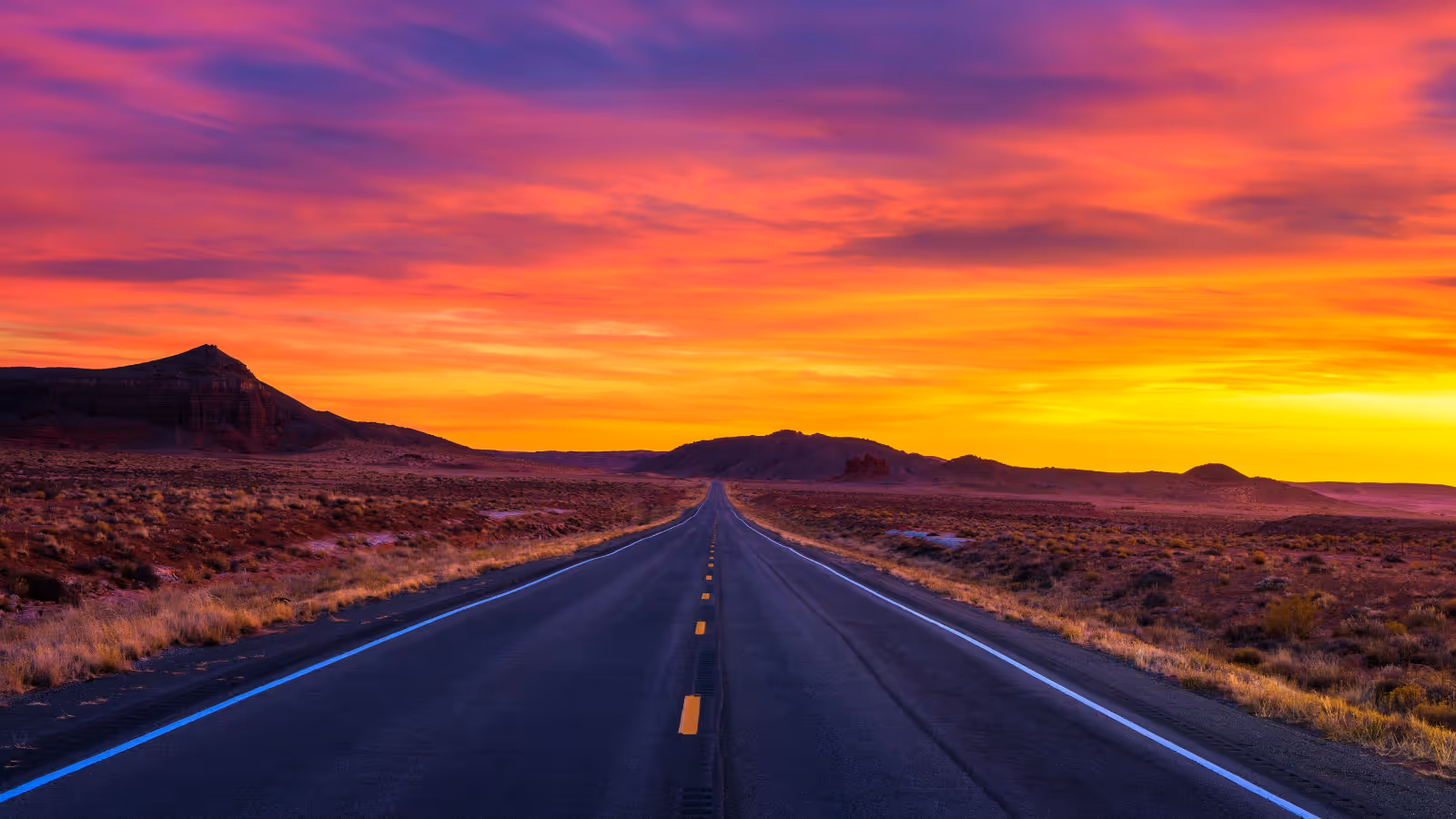 Empty highway stretches into desert landscape under a vibrant orange and purple sunset sky.