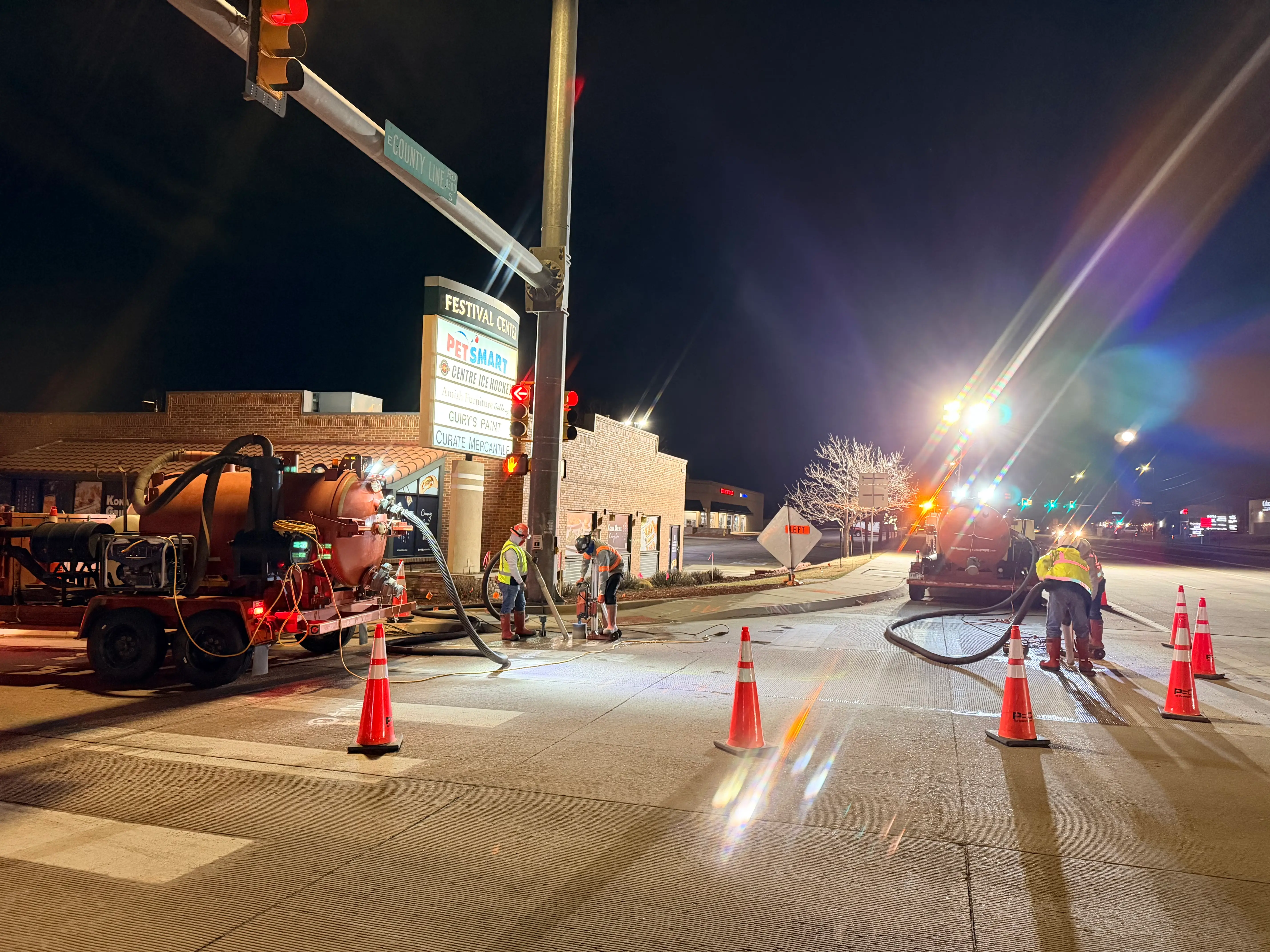 Nighttime roadwork scene with workers using machinery and cones blocking off a street near a lit Festival Center sign and a red traffic light.