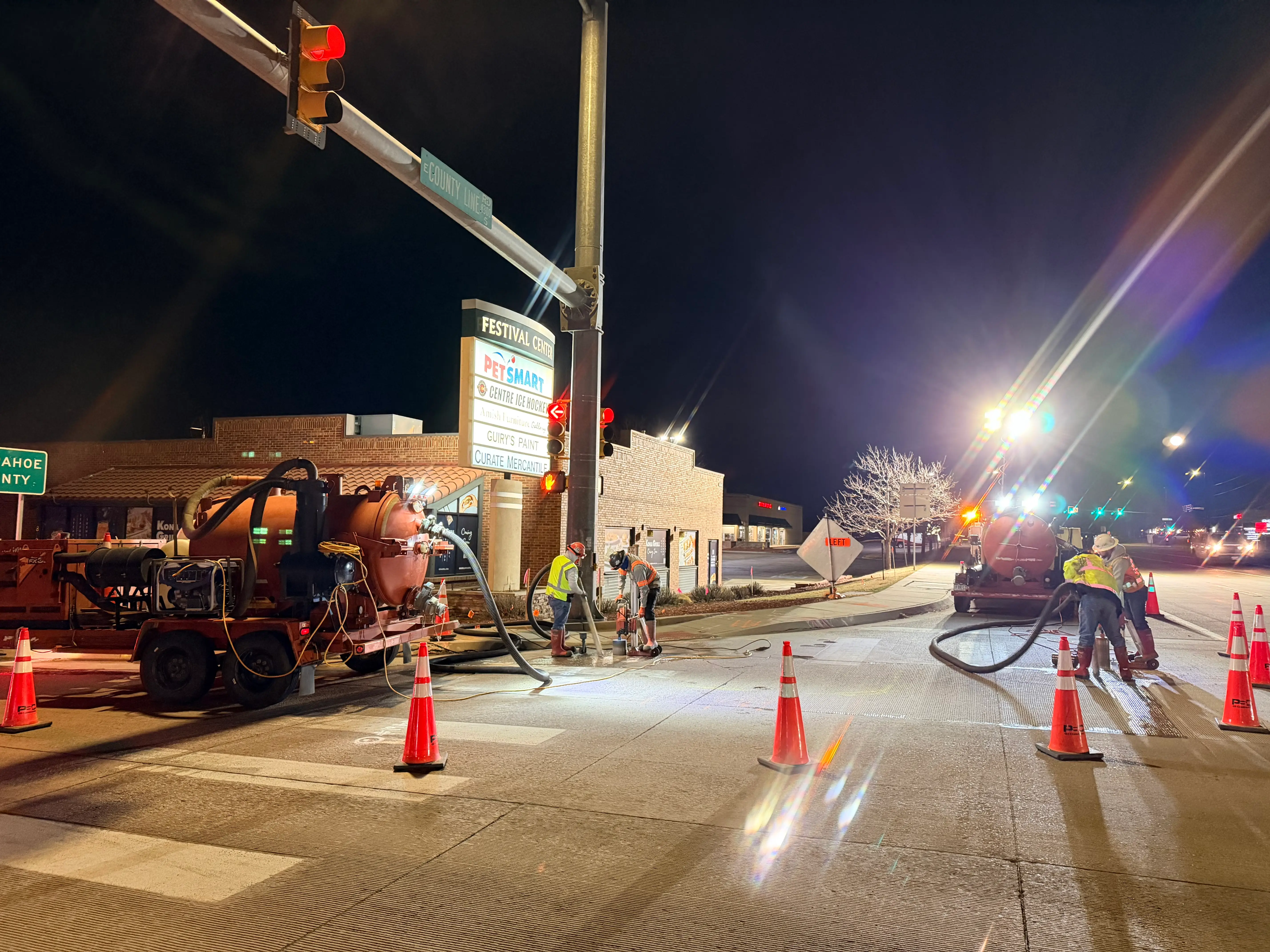 Nighttime roadwork scene with workers using machinery and cones blocking off a street near a lit Festival Center sign and a red traffic light.