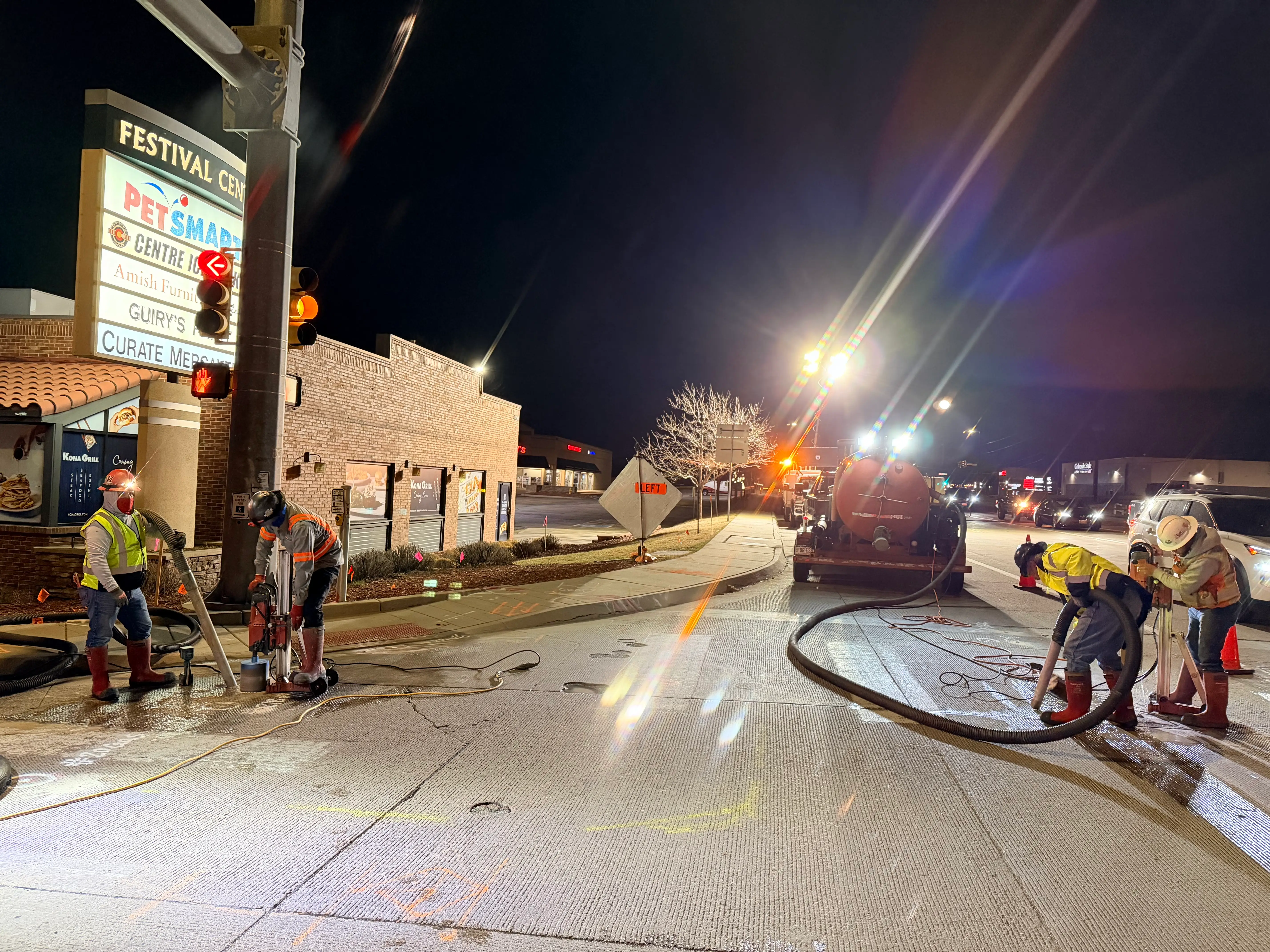 Nighttime roadwork scene with workers using machinery and cones blocking off a street near a lit Festival Center sign and a red traffic light.