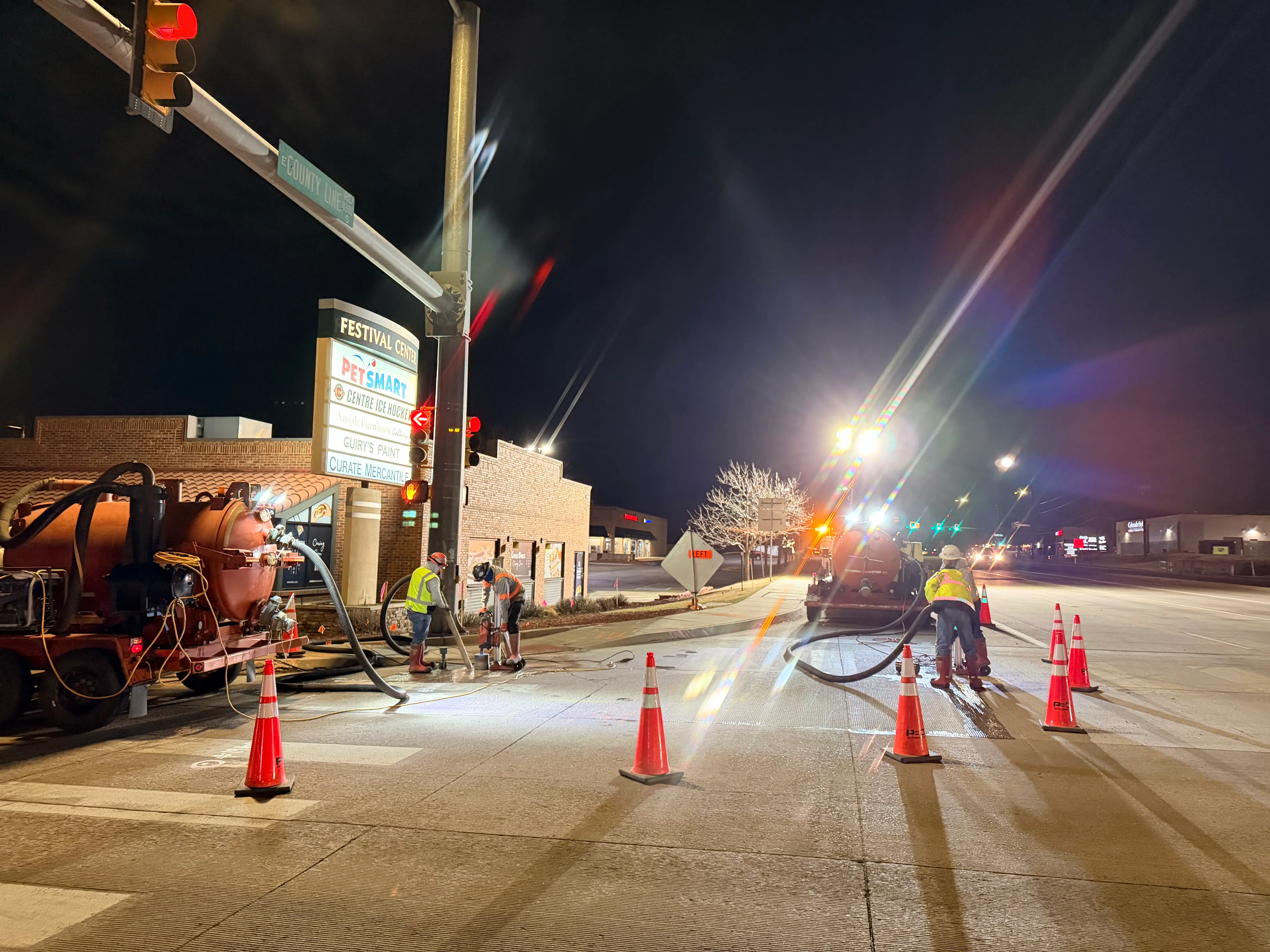 Nighttime roadwork scene with workers using machinery and cones blocking off a street near a lit Festival Center sign and a red traffic light.