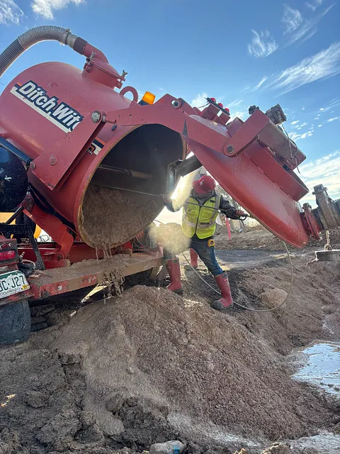 Nighttime roadwork scene with workers using machinery and cones blocking off a street near a lit Festival Center sign and a red traffic light.