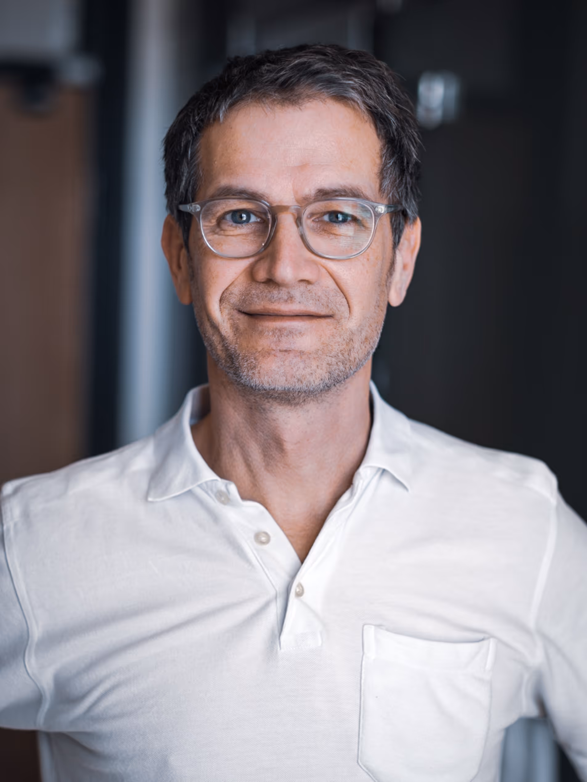 Mittelalterlicher Mann mit Brille lächelt in weißem Hemd vor dunklem Hintergrund.