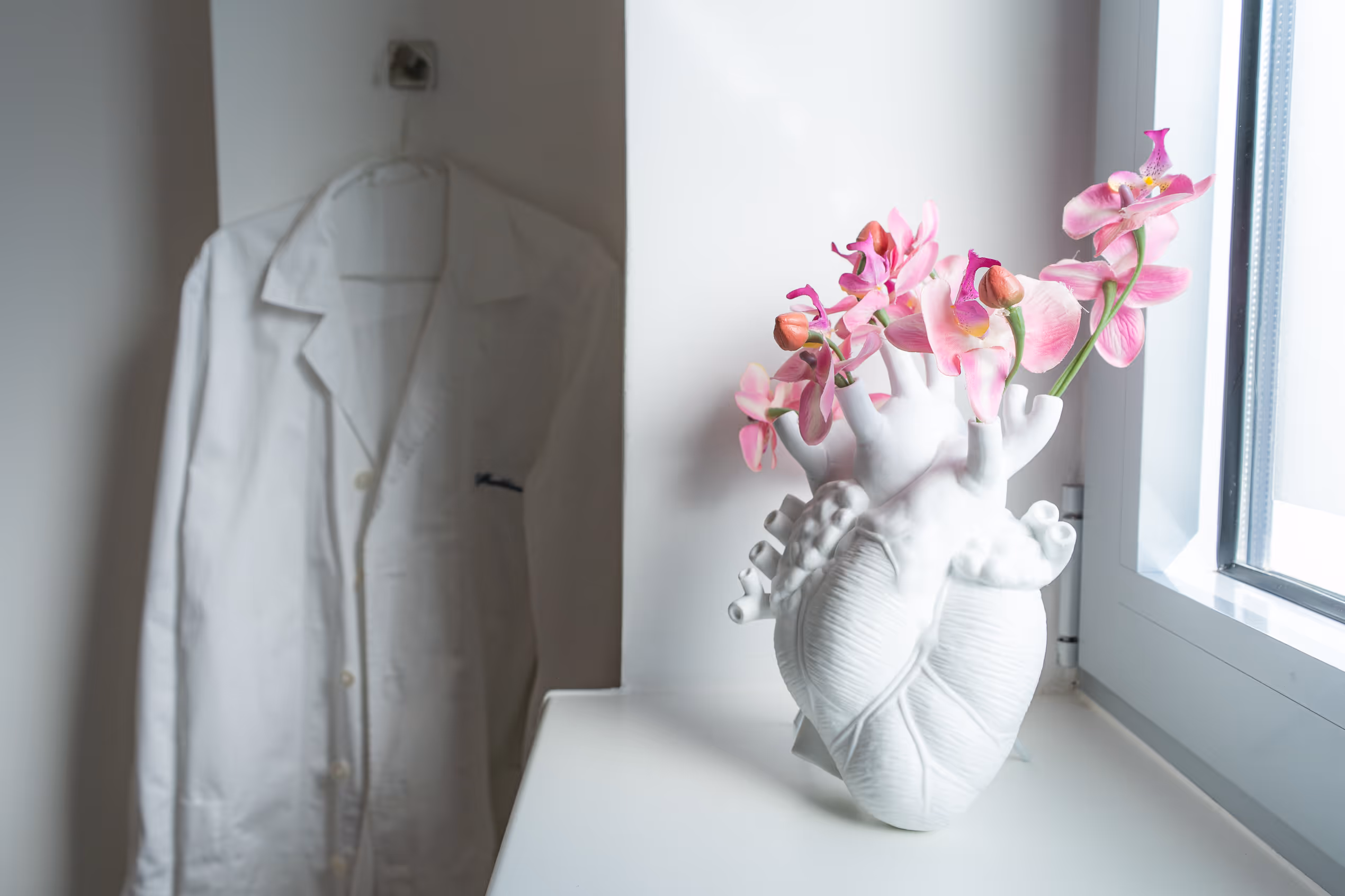 Blumenstrauß mit pinken Orchideen in weißem Herzvase neben weißem Laborkittel am Fenster.