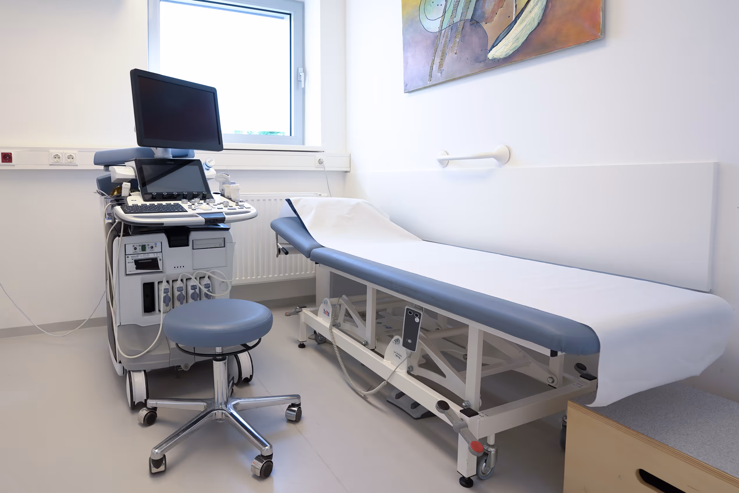 Moderne Untersuchungszimmer mit Ultraschallgerät, Untersuchungsbank und Rollstuhlstuhl.