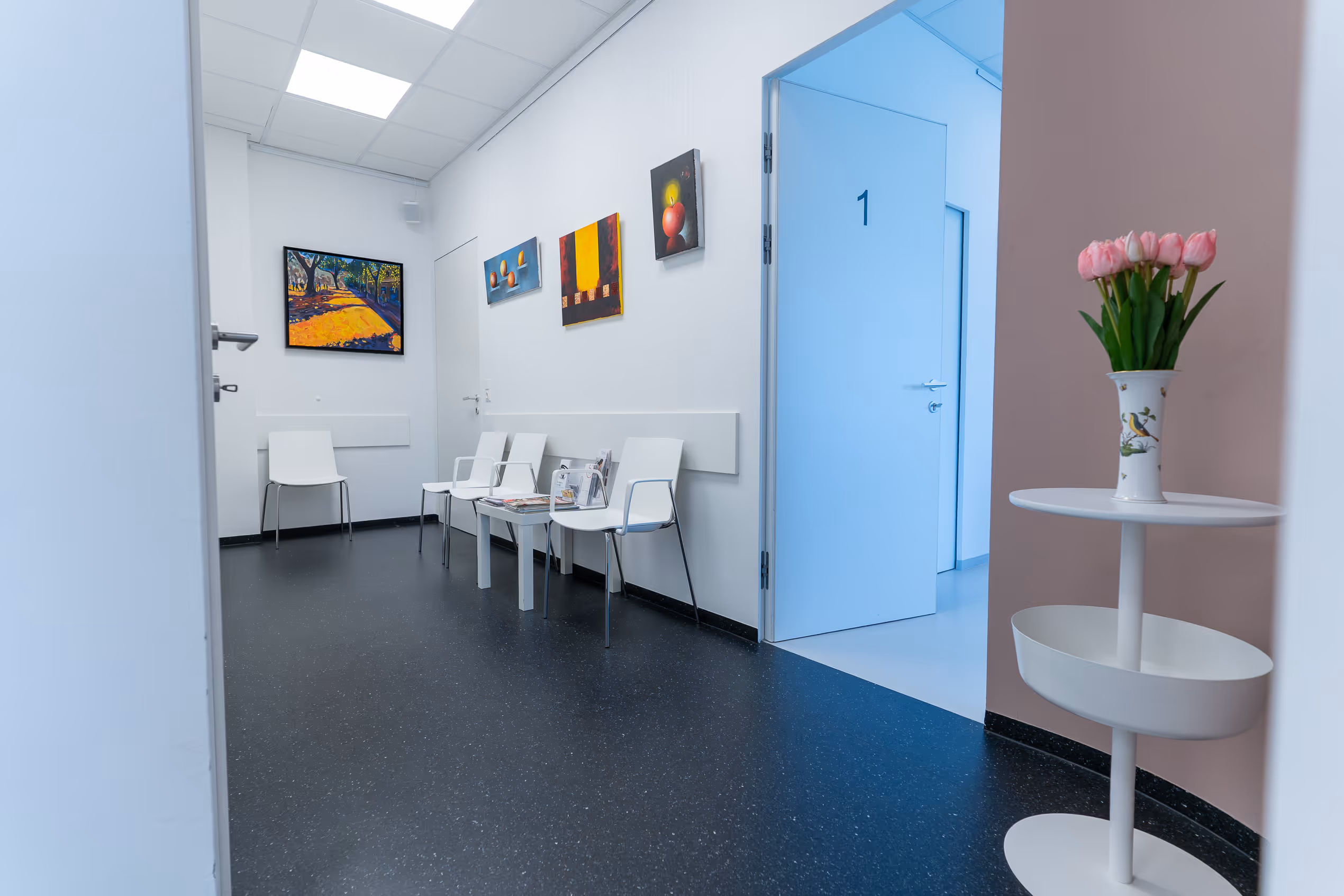 Wartezimmer mit weißen Stühlen, bunten Gemälden an der Wand und Vase mit rosa Tulpen.