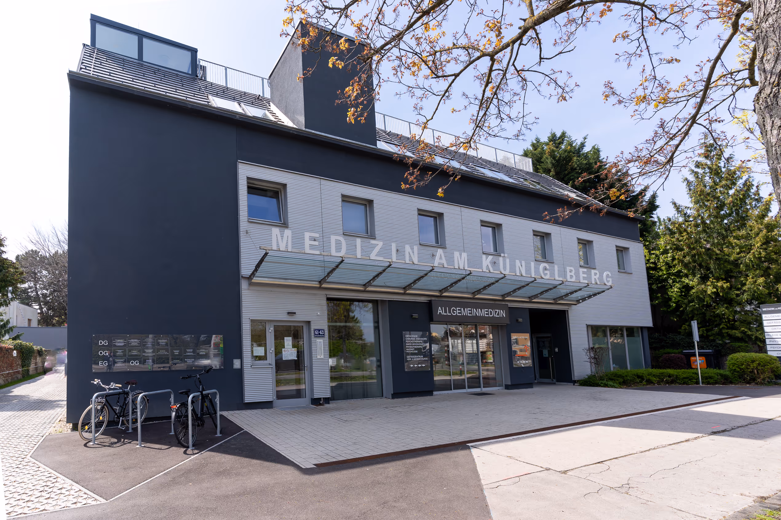 Modernes medizinisches Gebäude „Medizin am Königsberg“ mit Fahrradständern und Bäumen im Frühling.