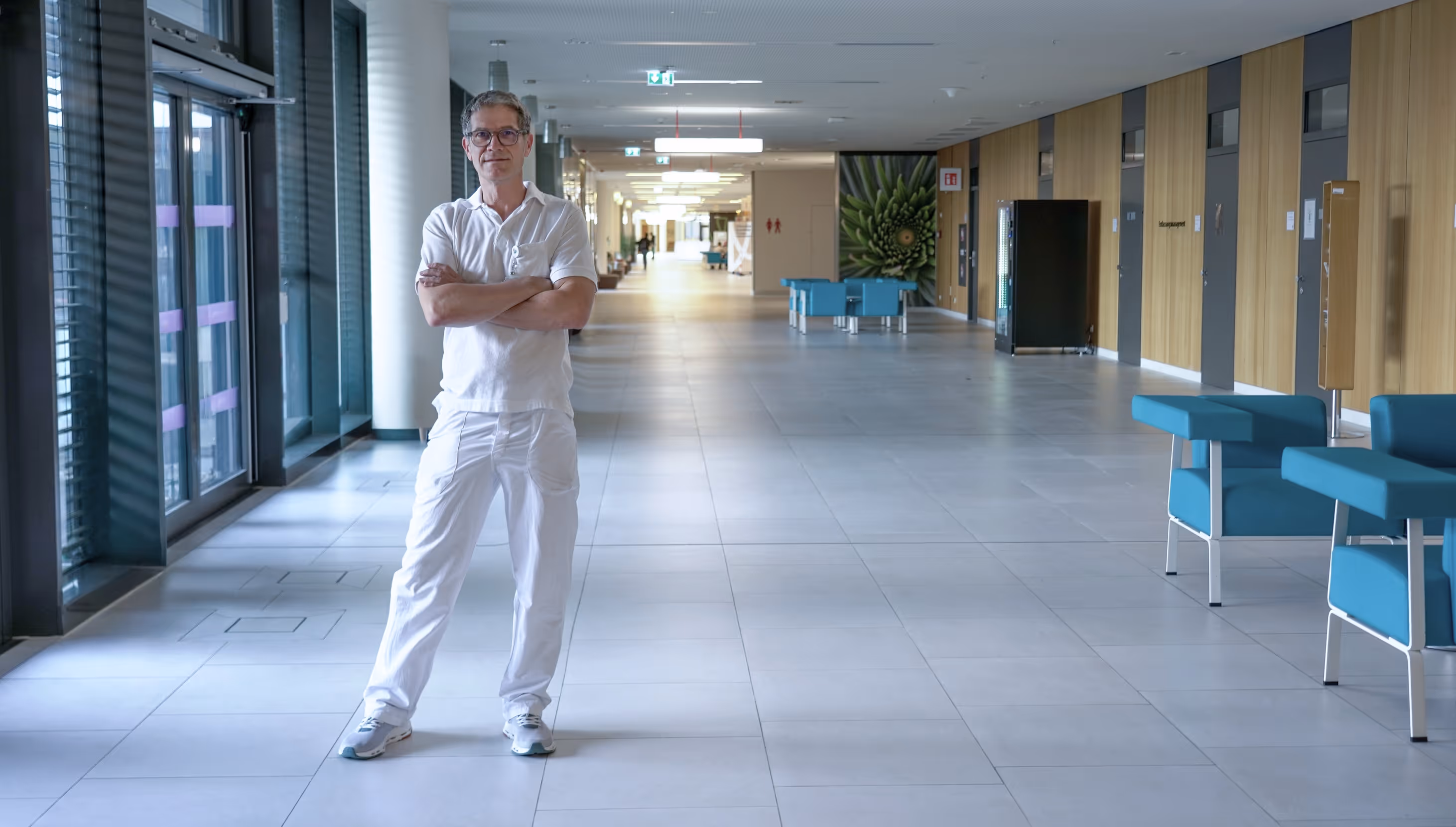 Dr. Gabriel Weiss in weißer Arbeitskleidung steht mit verschränkten Armen in einem hellen Gang.