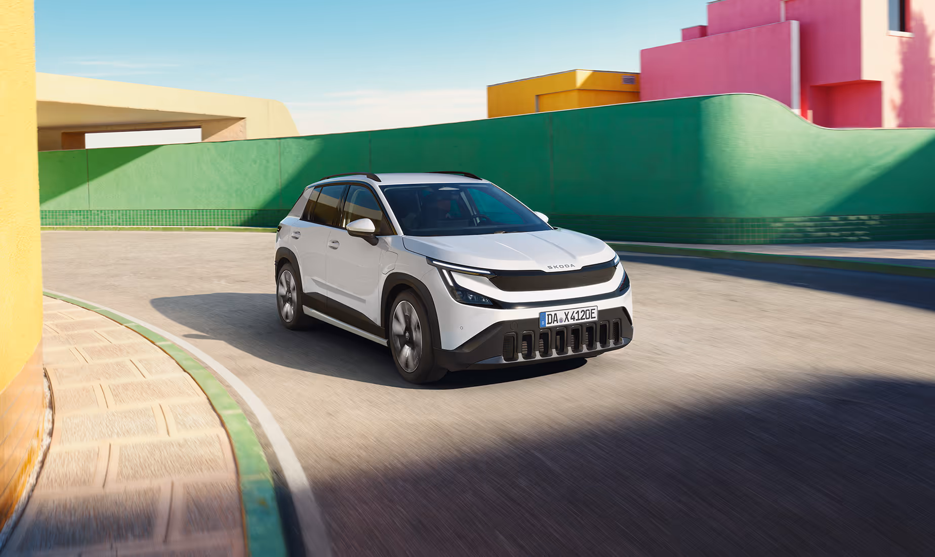 Ein Škoda Epiq in der Lackierung Platinum-Weiß Metallic fährt auf einer asphaltierten Straße. Das kompakte SUV-Crossover zeigt markante Designmerkmale wie dunkle Stoßfänger und eine geschlossene, schwarze Frontpartie. Im sonnigen Hintergrund ist eine moderne Architektur mit farbenfrohen, abstrakten Gebäudewänden in Grün, Gelb und Pink zu sehen.