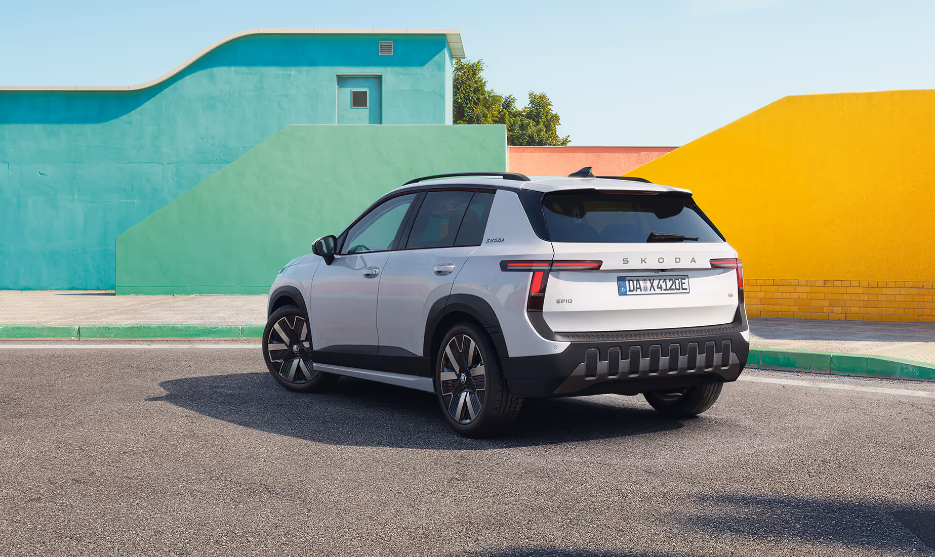Ein geparkter Škoda Epiq in der Lackierung Platinum-Weiß Metallic steht auf einer asphaltierten Fläche. Die Ansicht von schräg hinten zeigt markante, T-förmige LED-Rückleuchten, auffällige Leichtmetallfelgen sowie einen robusten, dunklen Stoßfänger mit vertikalen Elementen. Im sonnigen Hintergrund bilden geometrische Gebäudewände einen farbenfrohen Kontrast zum Fahrzeug.