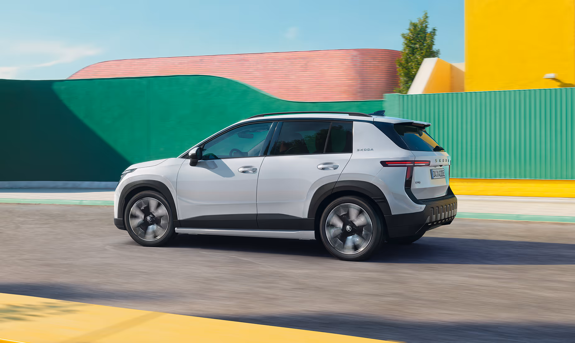 Ein Škoda Epiq in der Lackierung Platinum-Weiß Metallic fährt auf einer asphaltierten Straße. Die dynamische Seitenansicht zeigt dunkle Kontrastelemente an den Radkästen und Seitenschwellern, markante T-förmige Rückleuchten sowie rotierende Leichtmetallfelgen, die die Bewegung unterstreichen. Im sonnigen Hintergrund zieht eine moderne Architektur mit farbenfrohen, geometrischen Gebäudewänden vorbei.