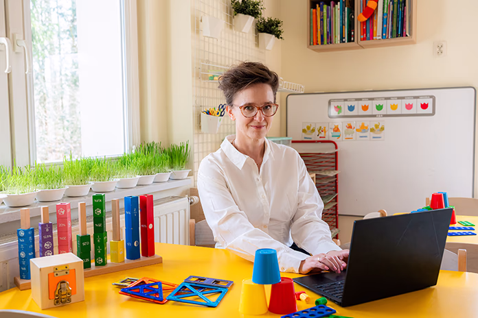 Kobieta w białej koszuli siedzi przy żółtym stole z laptopem i edukacyjnymi zabawkami w jasnym pokoju.