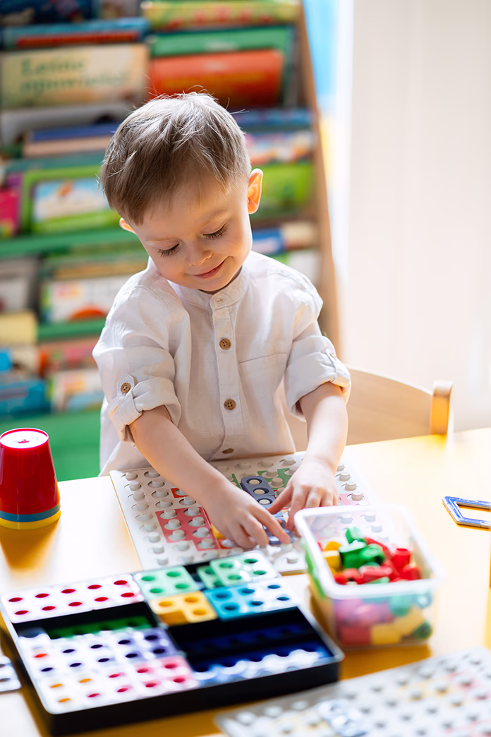 Mały chłopiec bawi się kolorowymi klockami edukacyjnymi przy stole, w tle regał z książkami.