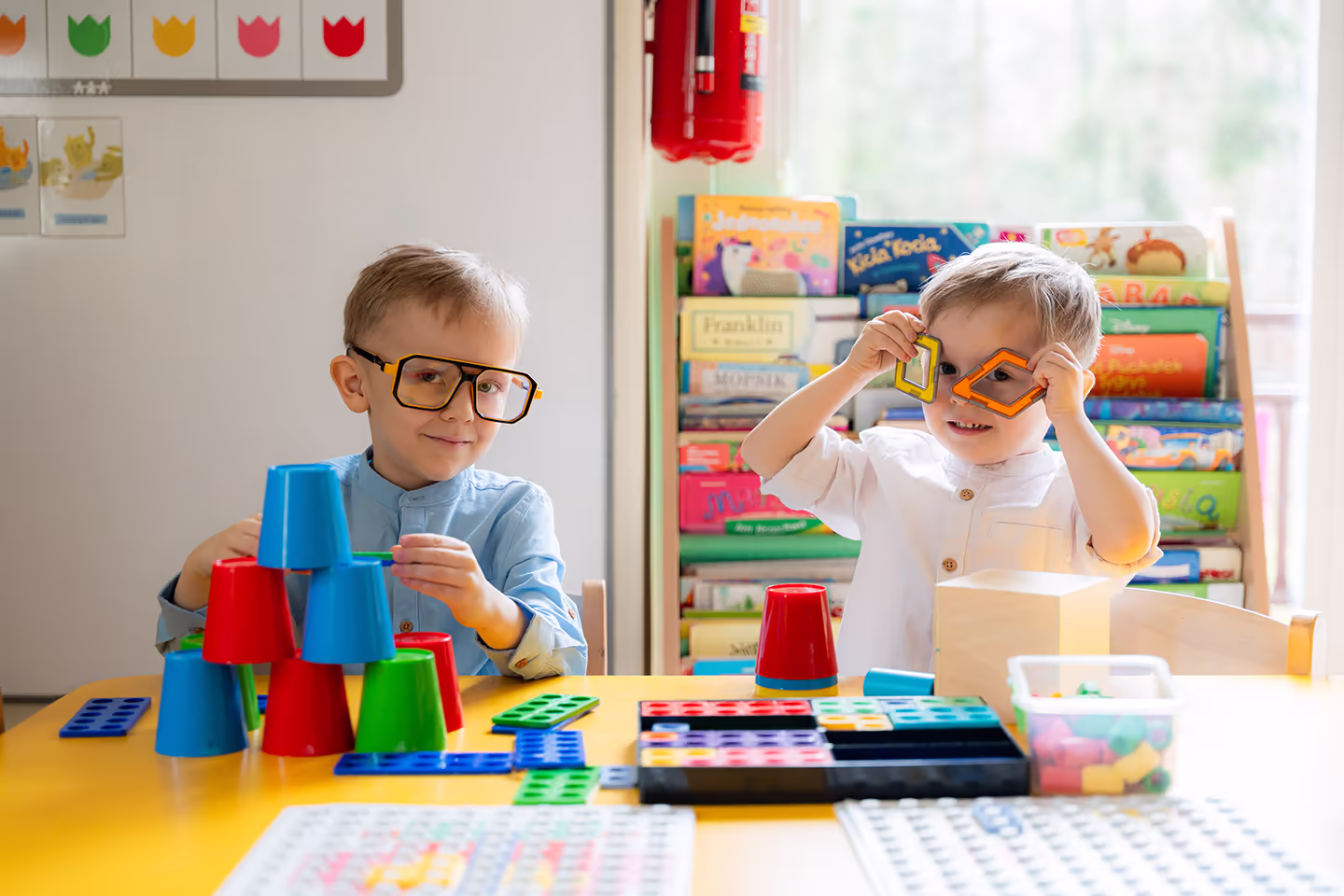 Dwóch chłopców bawiących się na stole kolorowymi kubeczkami i klockami edukacyjnymi w jasnym pokoju.