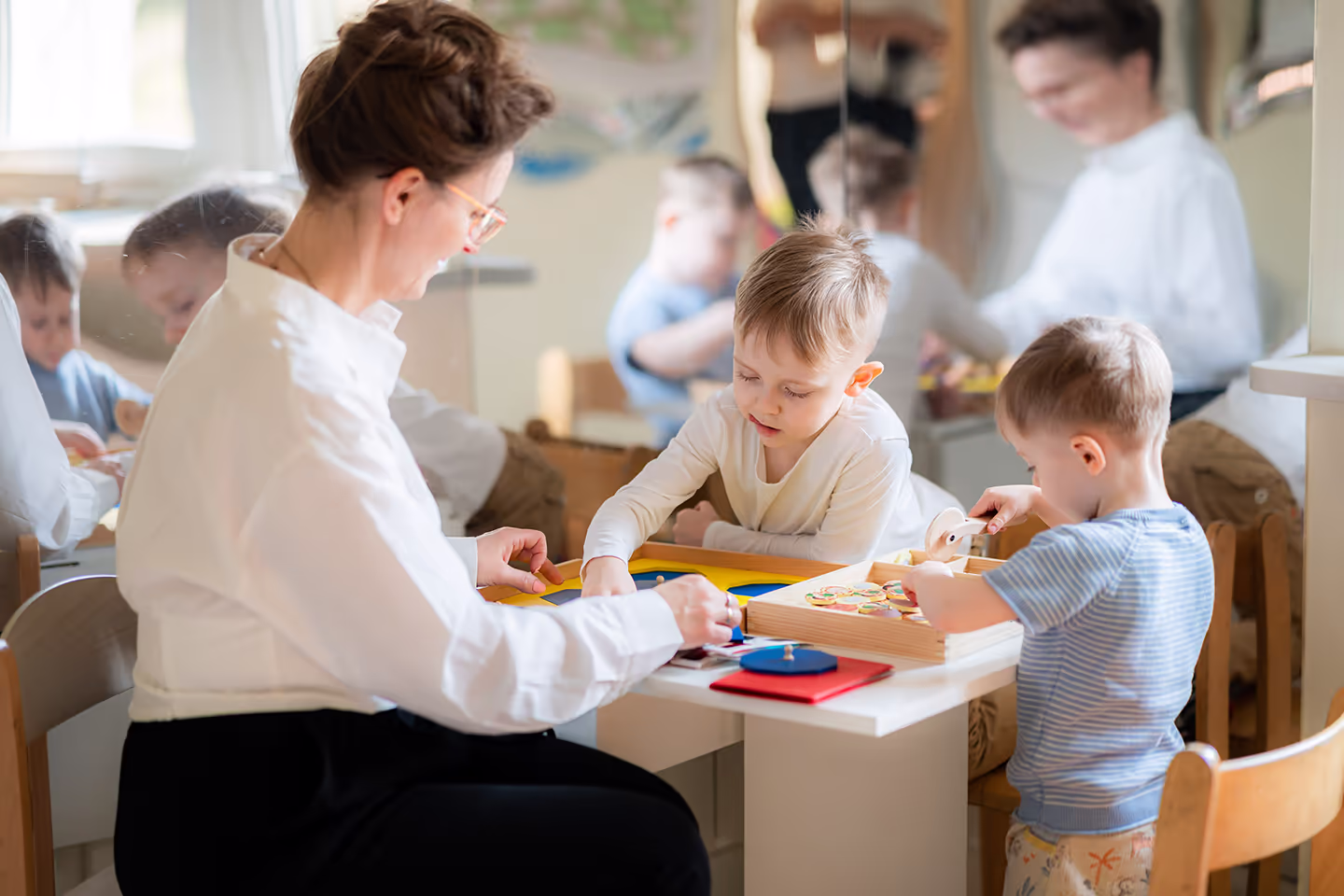 Kobieta wraz z dwójką małych chłopców bawią się edukacyjnymi zabawkami przy stole w jasnym pokoju.