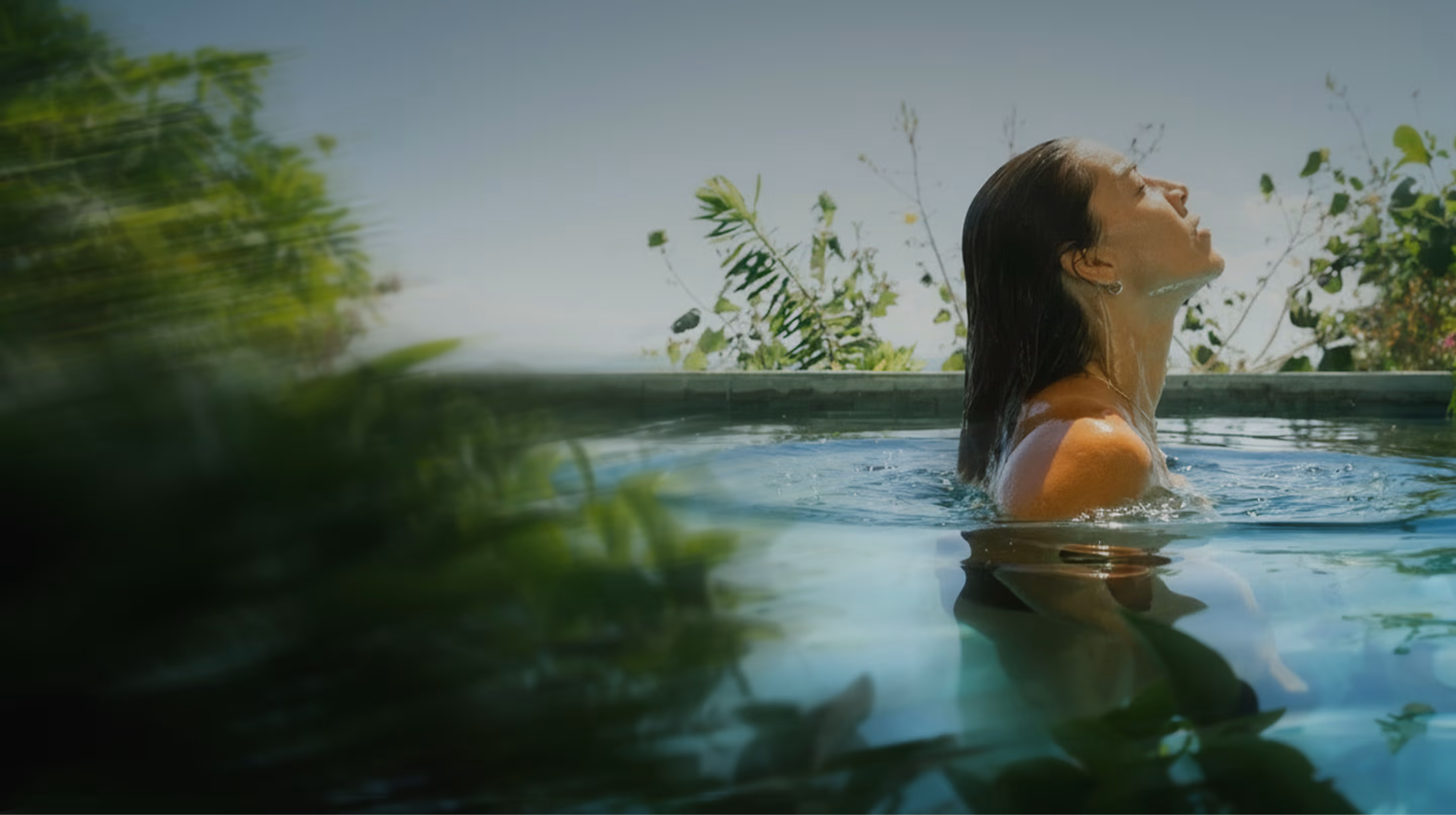 Woman with wet hair enjoying a peaceful moment in a pool surrounded by plants under clear sky.