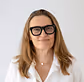 Woman with medium-length blonde hair wearing black glasses and a white blouse, smiling against a white background.