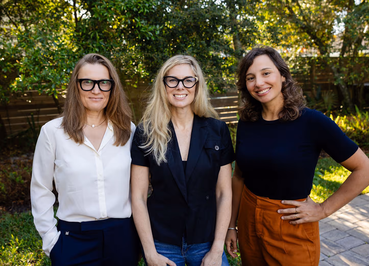 Three women standing outdoors smiling, with green foliage in the background.