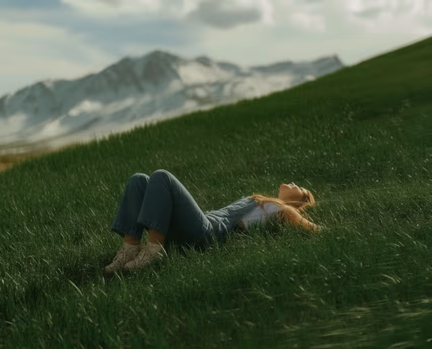 Woman lying on green grass on a hillside with mountains in the background.