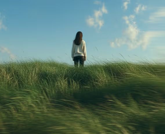 Person in white sweater standing in tall grass under a blue sky with scattered clouds.