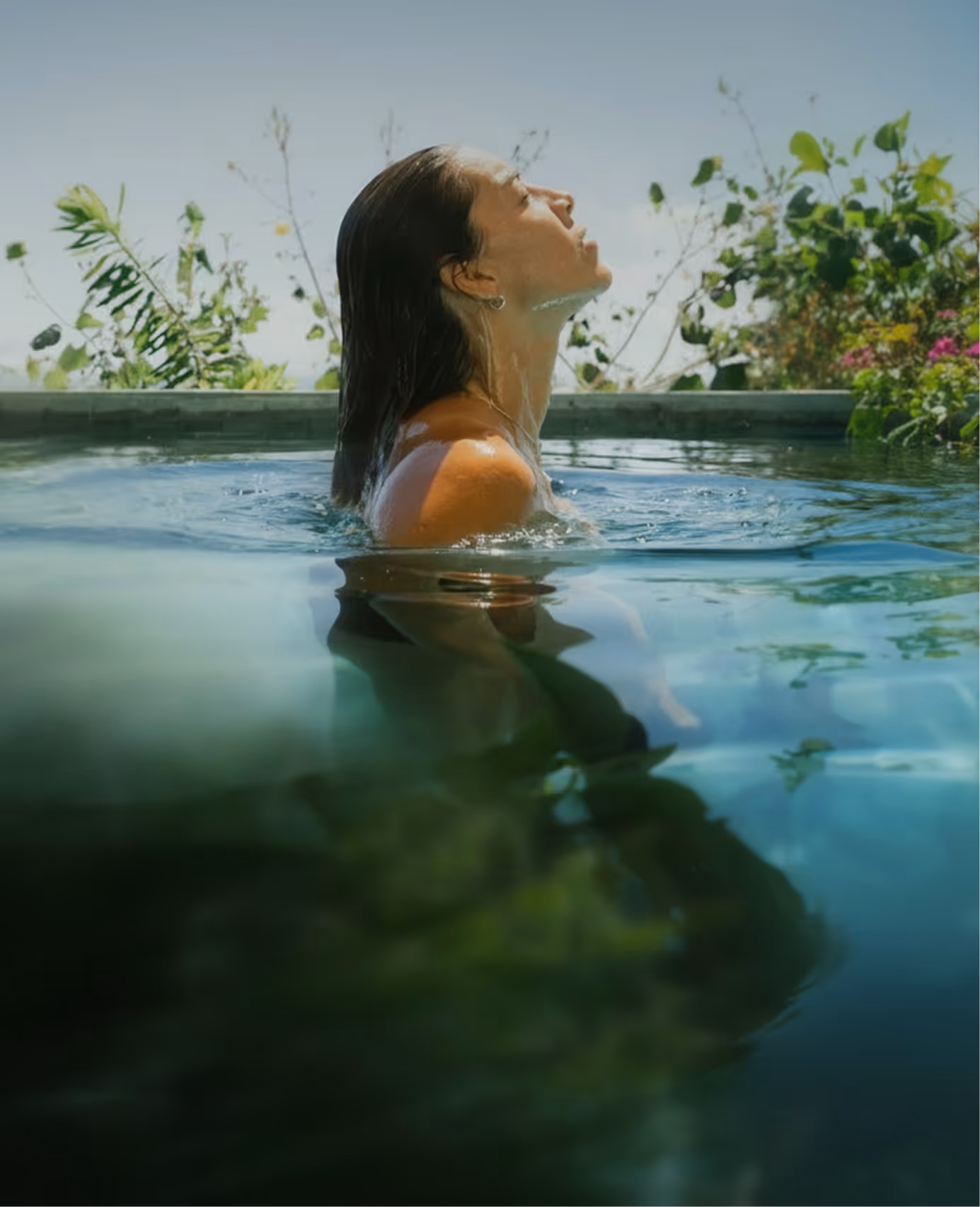 Woman with wet hair emerging from a pool surrounded by greenery in daylight.