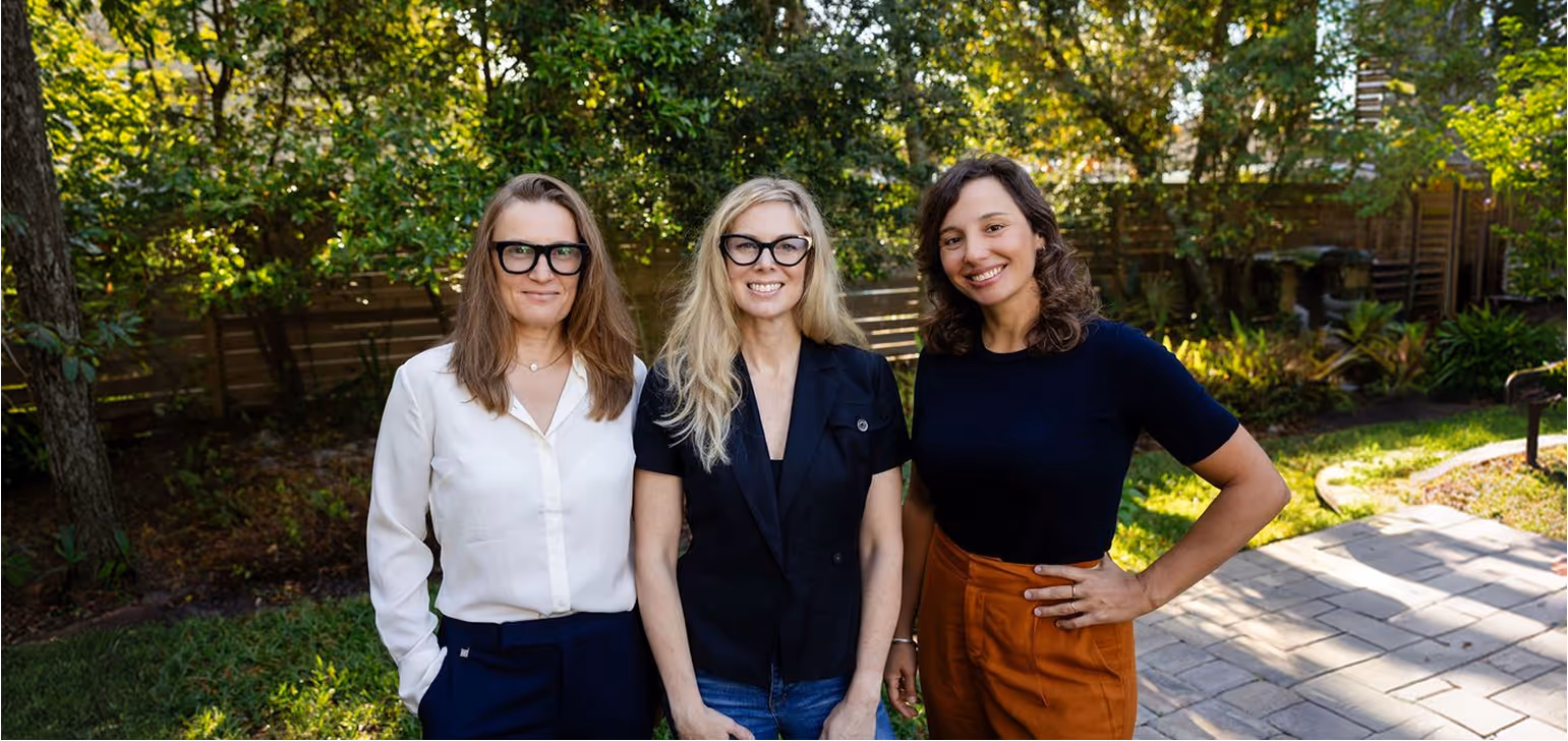 Three women standing side by side outdoors in a garden, smiling at the camera.