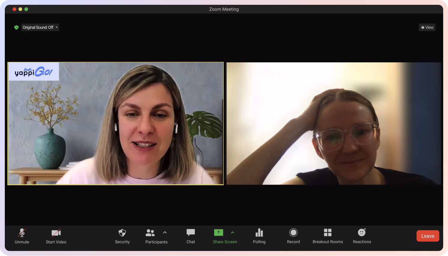 Zoom meeting with two women, one wearing earbuds and a pink shirt, the other with glasses and hand on head.