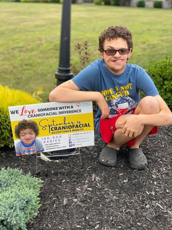 A young man holds a sign supporting craniofacial acceptance outdoors