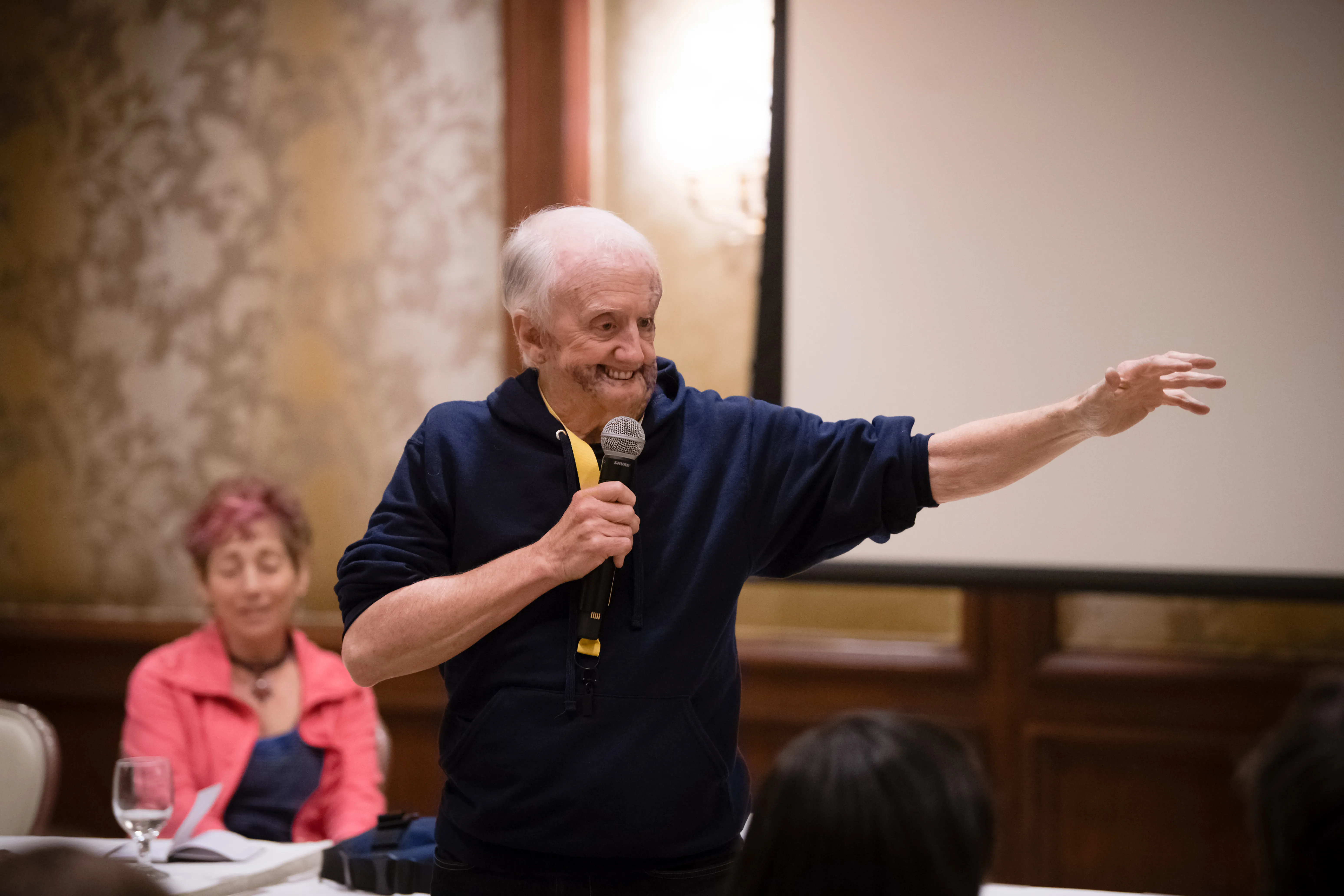 David Roche speaks with a microphone, arm outstretched, to an audience