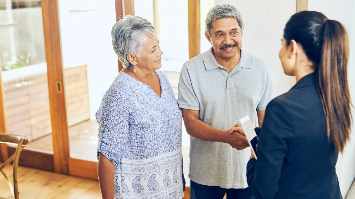 Older couple shaking hands with a woman holding a tablet in a bright room.