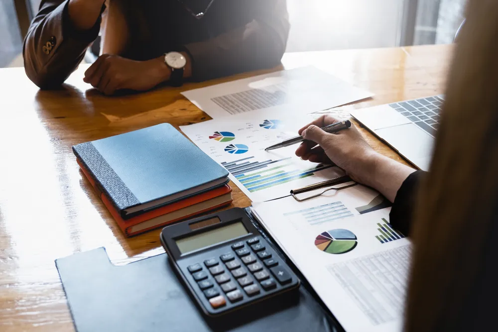 Two people discussing charts and graphs on paper at a wooden desk with a calculator and notebooks.