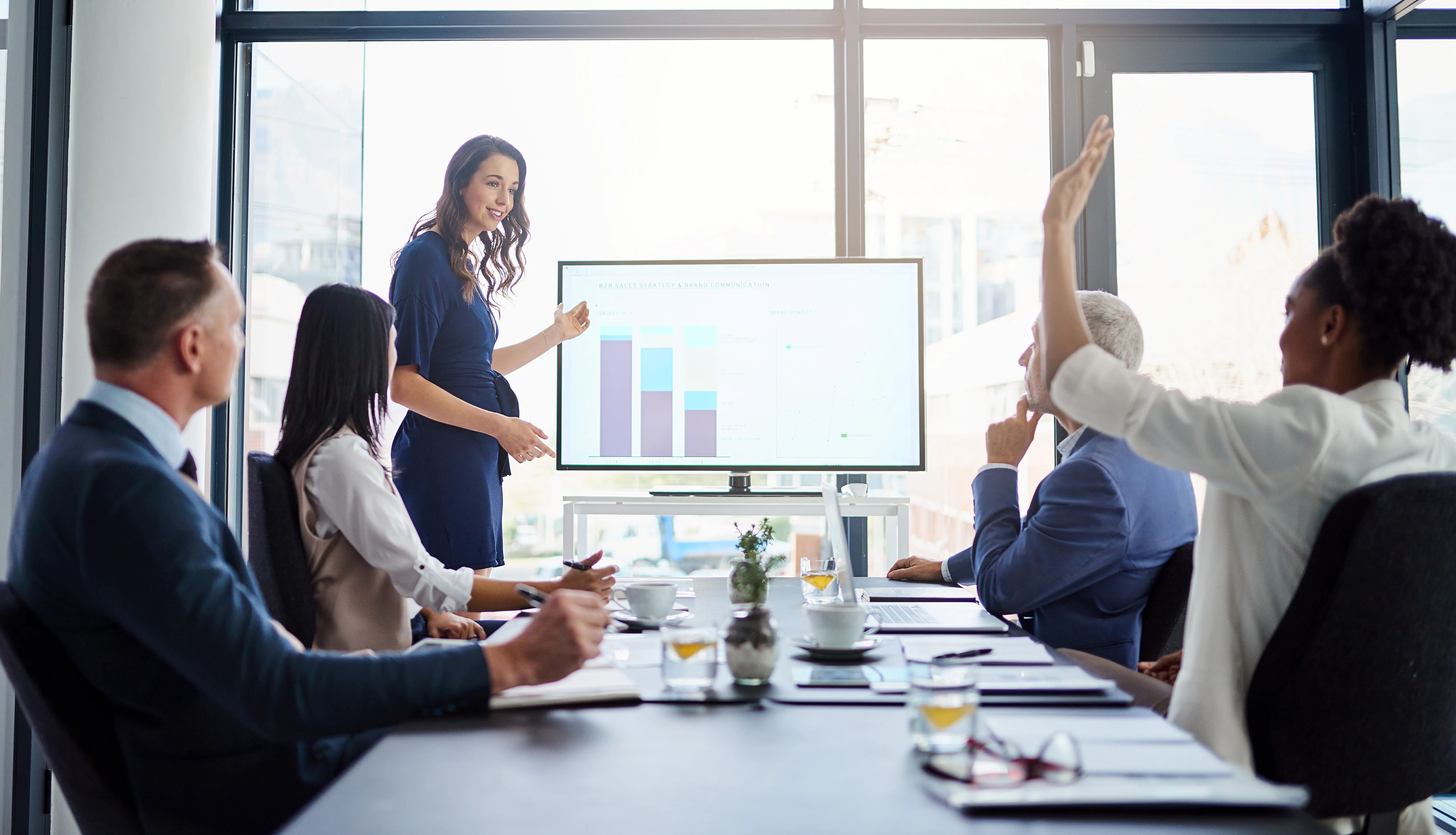 Woman giving a presentation to colleagues in a bright conference room with charts on screen.