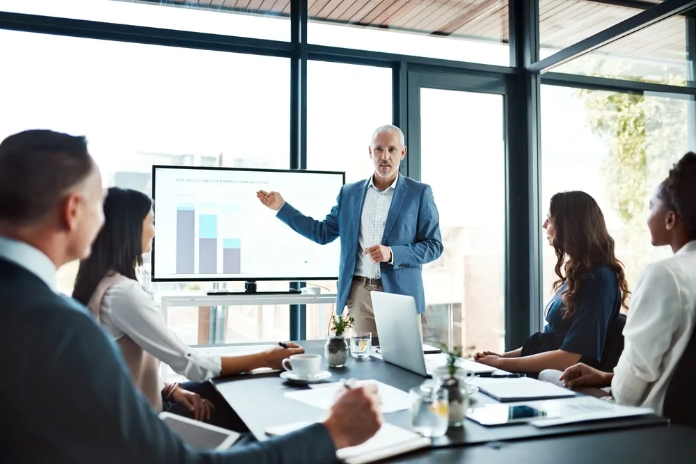 Businessman in a blue suit giving a presentation to four colleagues in a bright meeting room.