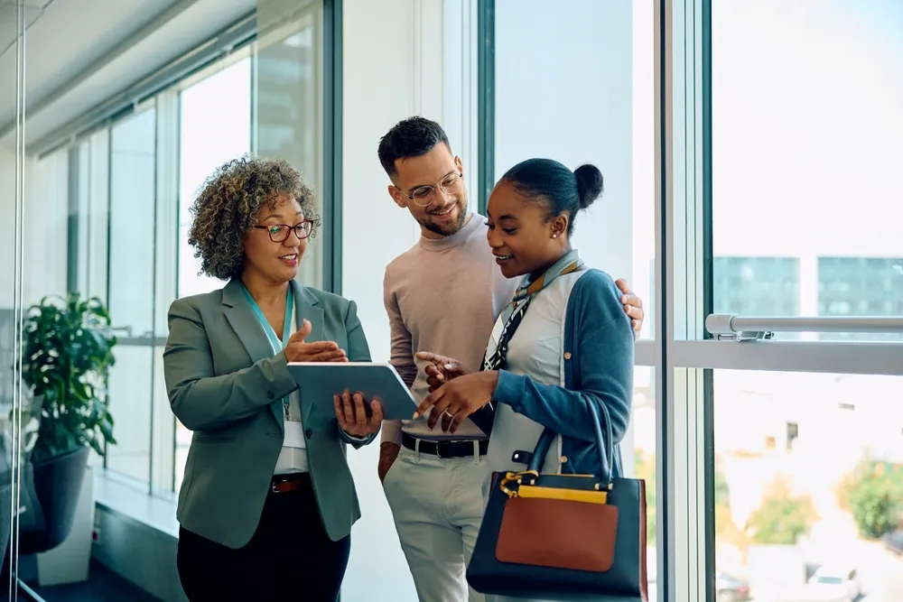 Three colleagues in office hallway discussing something on a tablet, smiling and engaged.