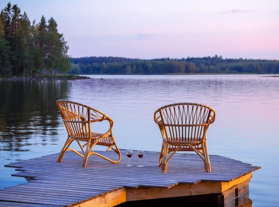 Two empty wicker chairs and two glasses of wine on a wooden dock by a calm lake at sunset.