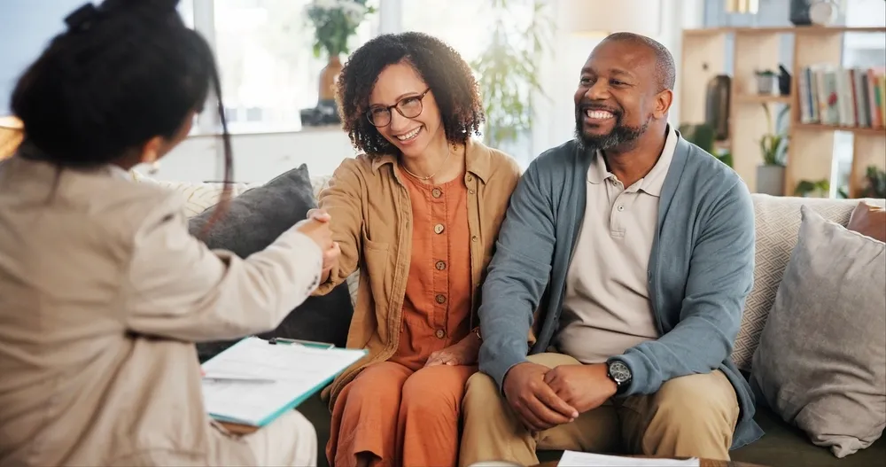 Smiling couple shaking hands with a professional across the couch in a cozy, bright room.