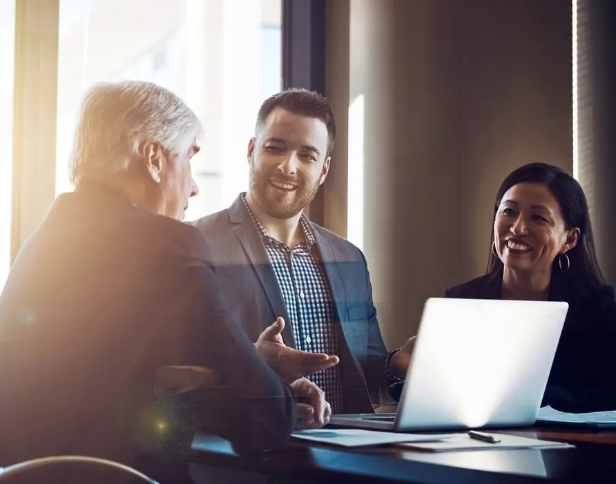 Three businesspeople in a meeting room with a laptop, engaged in a friendly discussion.