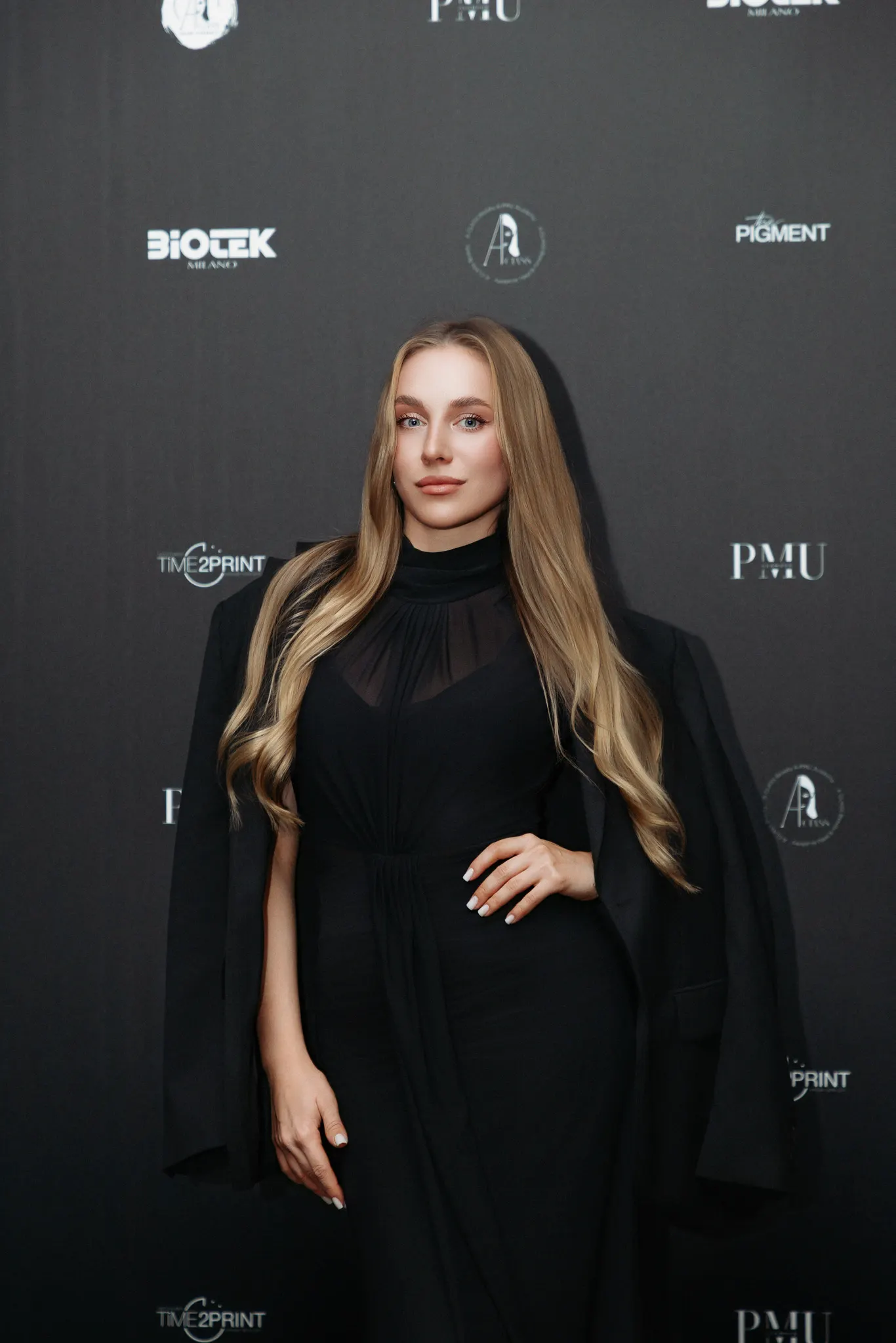 Blonde woman with long hair in a black dress posing before a black backdrop with logos.