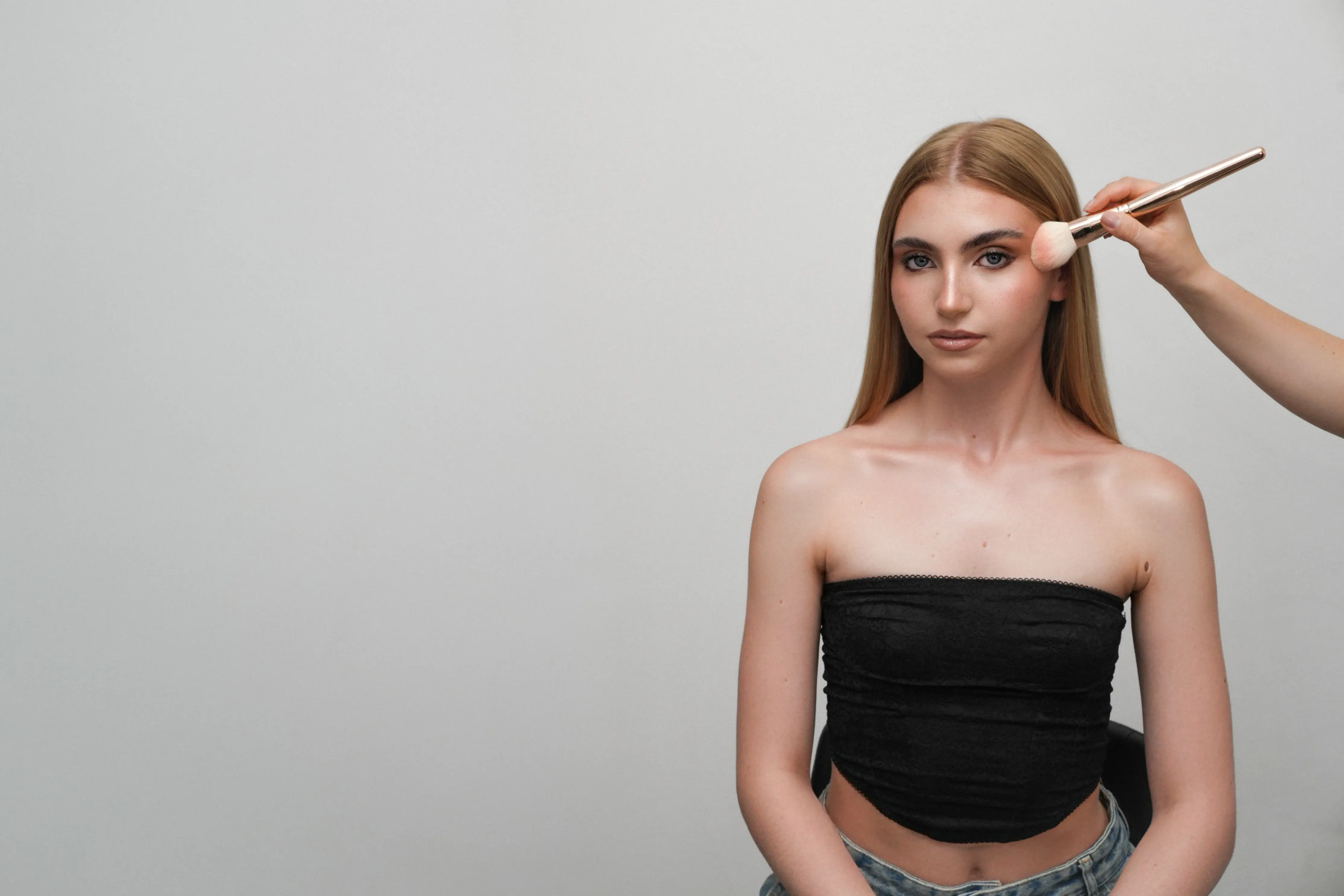 Woman with long blonde hair and black tube top having makeup applied with a brush.