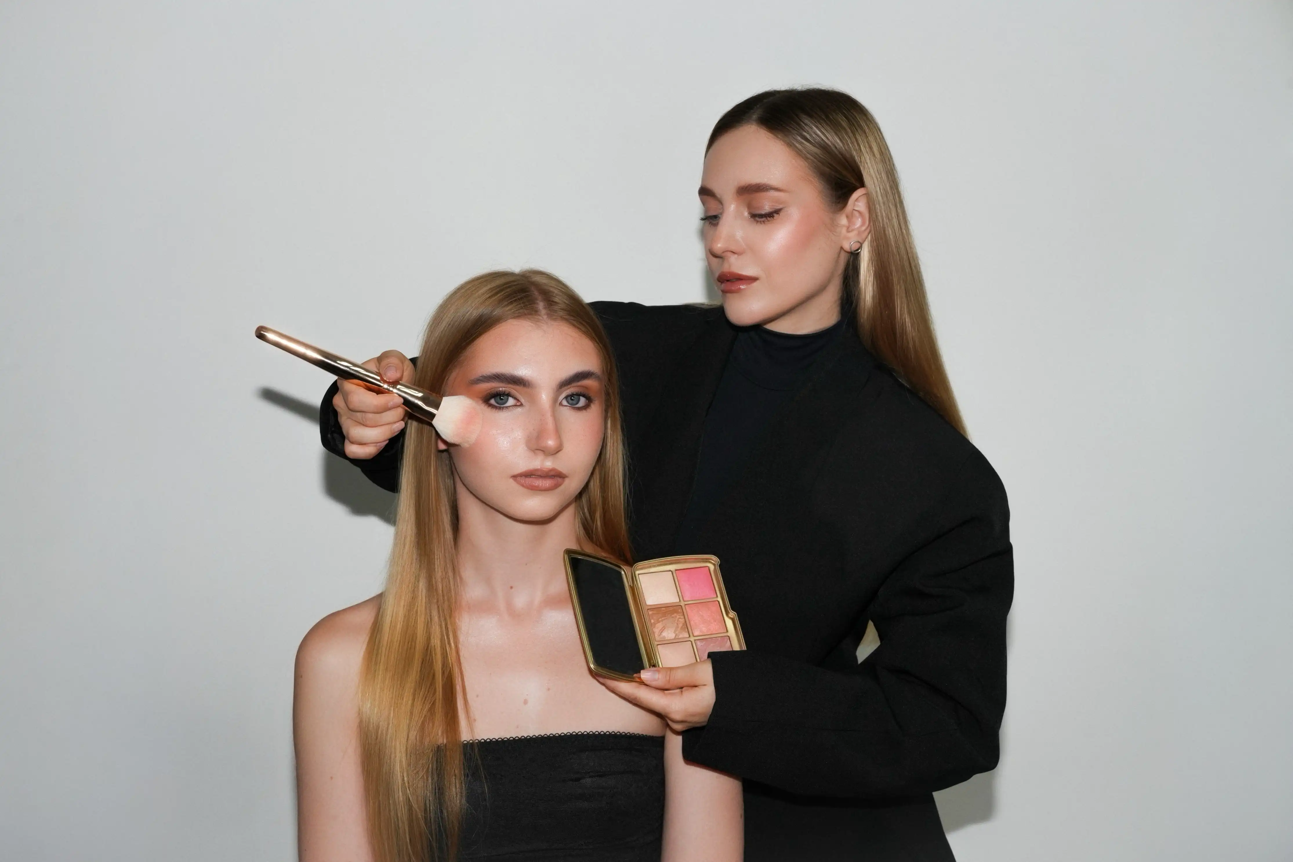 Woman applies makeup blush with brush on another woman's cheek while holding a palette with blush shades.