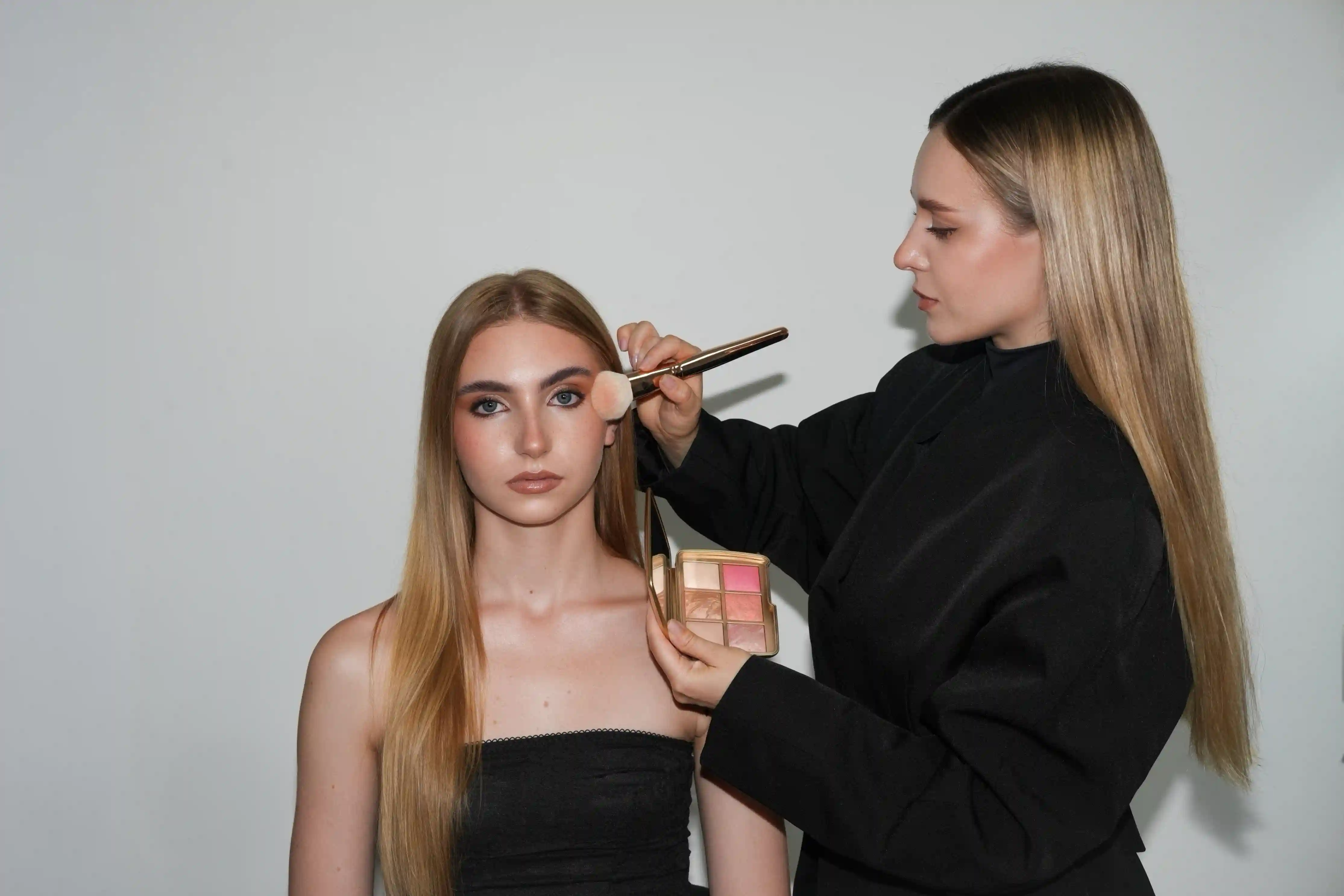 Makeup artist applying blush with a brush to a seated young woman with long blonde hair.