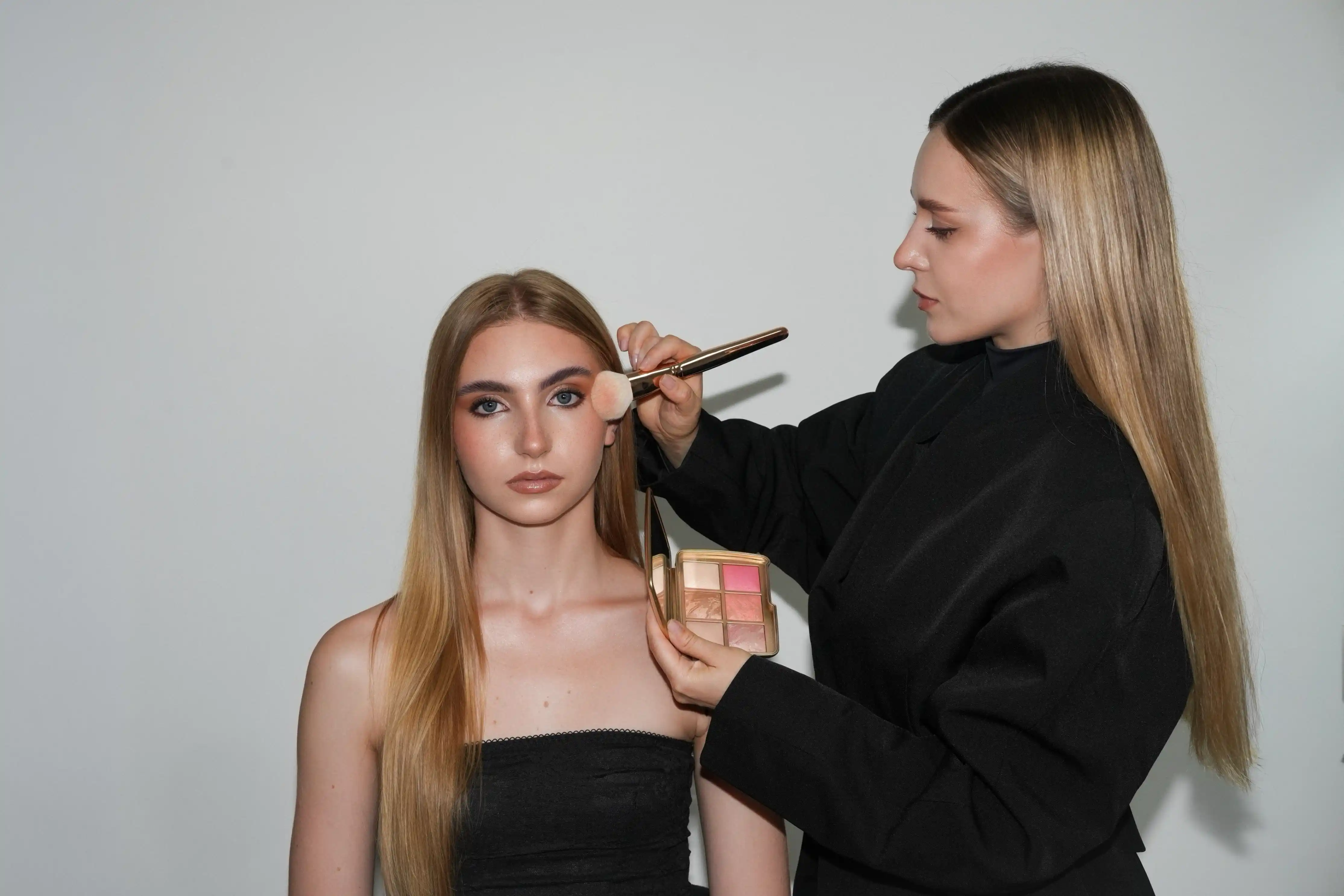 Makeup artist applying blush with a brush to a seated young woman with long blonde hair.