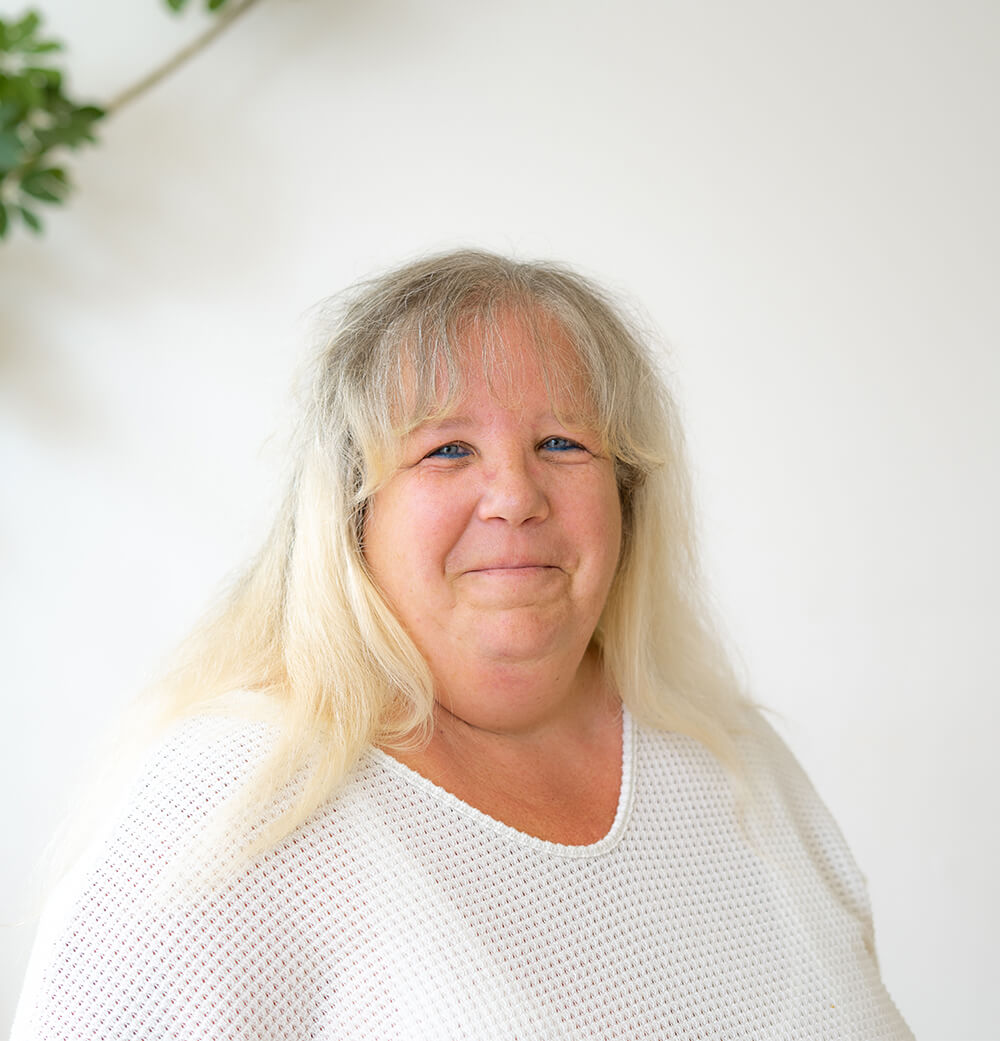 Lächelnde Frau mittleren Alters mit langem blonden Haar und weißem Pullover vor unscharfem Hintergrund.