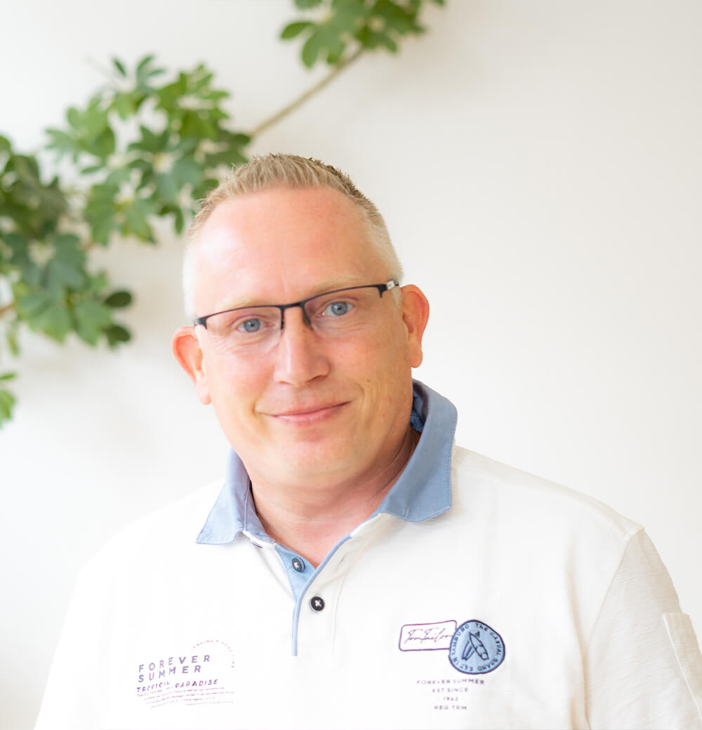 Ein lächelnder Mann mit kurzem blonden Haar und Brille, der ein weißes Polohemd mit blauem Kragen trägt; ein rosa Oberteil und ein schwarzer Blazer vor weißem Hintergrund.