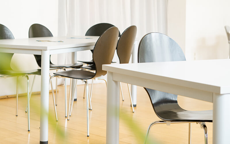 Leeres Klassenzimmer mit weißen Tischen und schwarzen, modernen Stühlen auf einem hellen Holzboden.