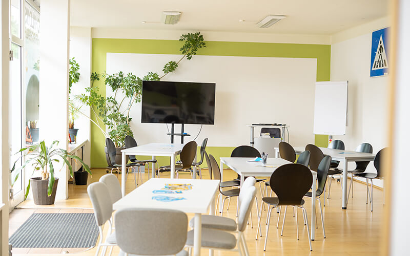Heller Unterrichtsraum mit Tischen, Stühlen, Fernsehbildschirm, Whiteboard und Pflanzen an den Fenstern.
