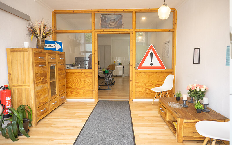Heller Raum mit Holzmöbeln, weißen Stühlen, Pflanzen und einem Warnschild an einer Trennwand.