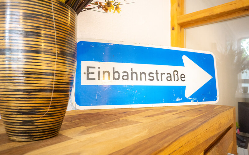 Blaues deutsches Einbahnstraßenschild mit weißem Pfeil auf einer Holzoberfläche neben einer gestreiften Vase mit Trockenblumen.